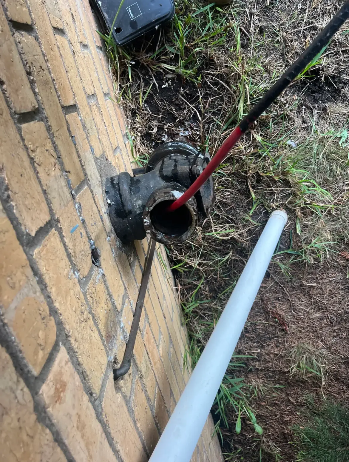 Black cast iron junction on a brick wall with a red jetter hose going in and a white pipe lying beside it