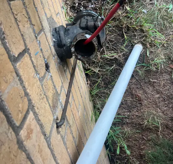 Black cast iron junction on a brick wall with a red jetter hose going in and a white pipe lying beside it