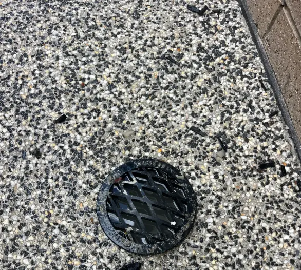 Black grate over a drain in exposed-aggregate paving beside a brick wall