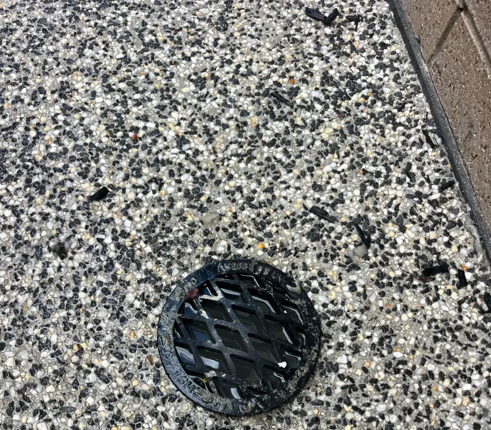 Black grate over a drain in exposed-aggregate paving beside a brick wall