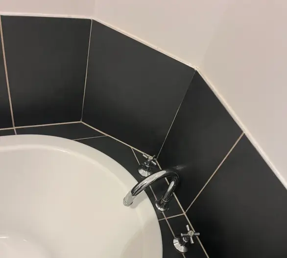 Corner bath with a chrome spout and cross-handle taps against black tiles