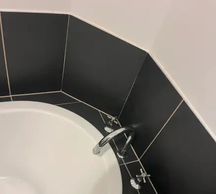 Corner bath with a chrome spout and cross-handle taps against black tiles