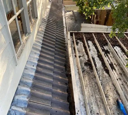 Debris-filled gutter between a tiled roof and a metal roof beside a wall