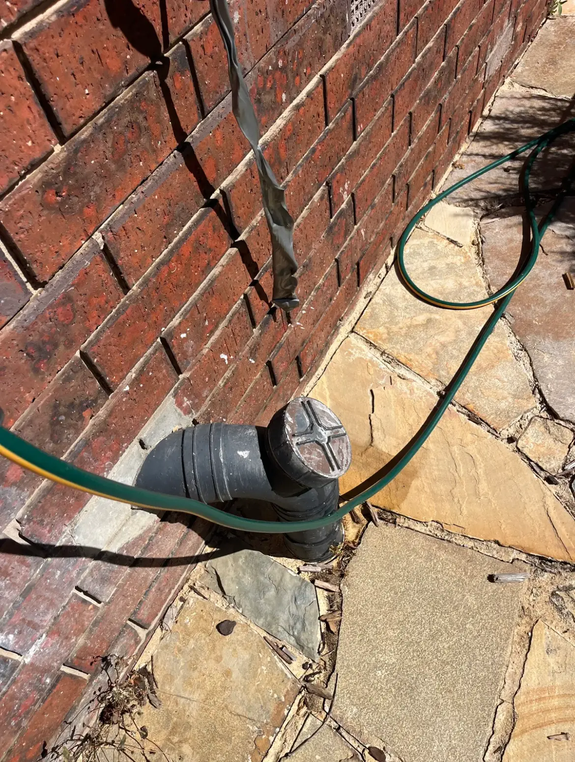 Drain cleanout point at a brick wall with a hose running to it on paving
