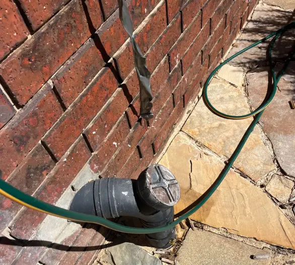 Drain cleanout point at a brick wall with a hose running to it on paving