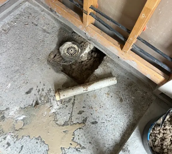 Exposed wall framing with black pipes and an opened floor drain on a concrete slab
