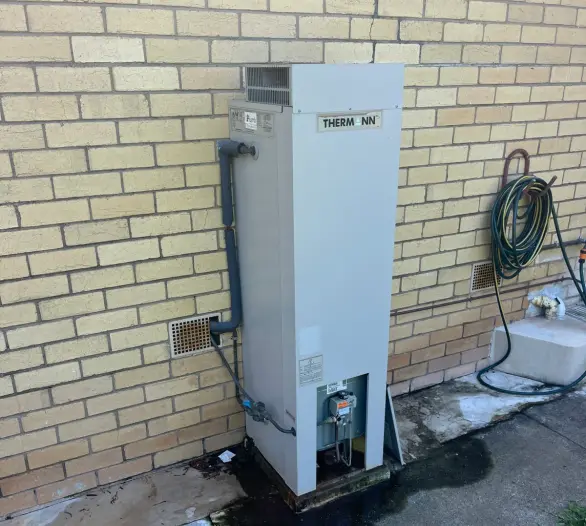 Gas storage hot water unit against a brick wall with wet staining on the ground beneath it