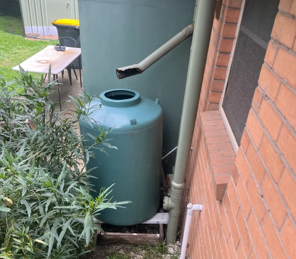 Green rainwater tank on a timber base next to a brick wall, with a downpipe and white overflow pipe