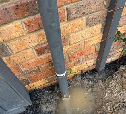 Grey downpipe at a brick wall entering a muddy dug hole, with tools on mulch