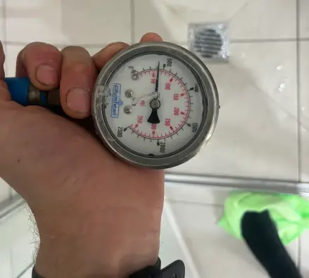 Hand holding a pressure gauge over a shower floor drain with a blue hose attached