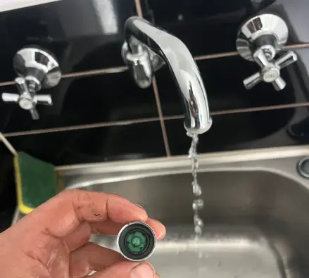 Hand holding a tap aerator beside a running spout over a stainless sink