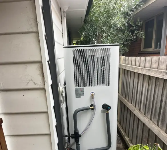 Heat pump hot water unit with insulated pipework beside a side fence