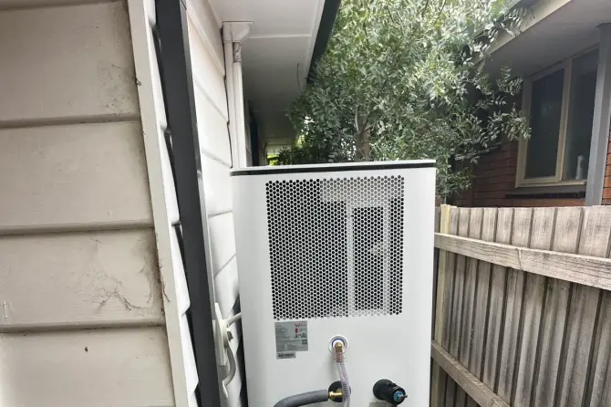 Heat pump hot water unit with insulated pipework beside a side fence