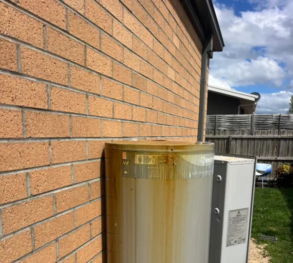 Hot water tank against a brick wall with rust streaking and wet staining under the pipe fittings