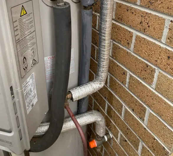 Insulated hot water pipework and relief valve on a hot water unit beside a brick wall
