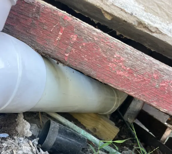 Large white pipe passing under a red-painted timber beam with a black cap and weeds below