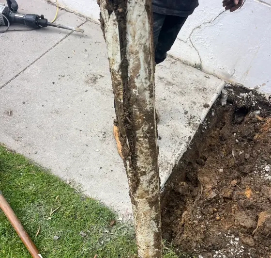 Muddy old pipe held upright beside a dug-out hole next to a path, with a breaker tool lying at the top left