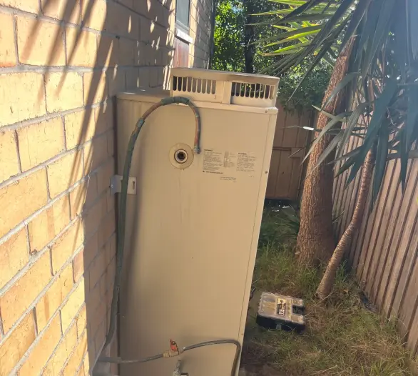 Older gas hot water unit with hoses and pipework against a wall in a narrow side passage
