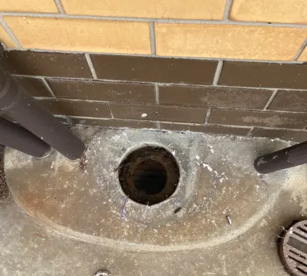 Open round drain in a curved concrete apron beneath two downpipes on a yellow brick wall, work boots at the bottom