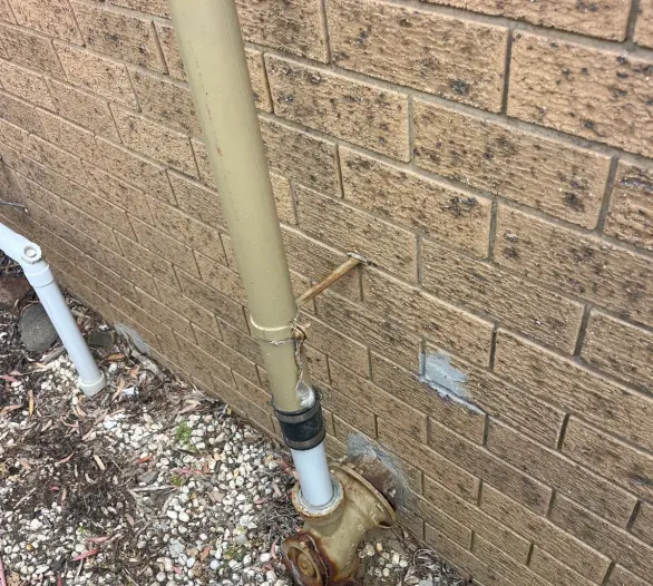 Painted downpipe and brass valve fitting at the base of a brick wall above white gravel