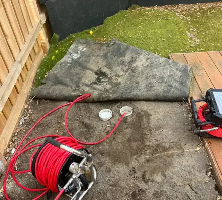 Red high-pressure hose reel and camera case beside two open drain caps in the soil under folded-back artificial turf