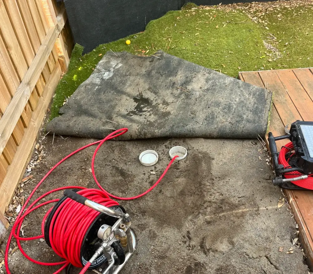 Red high-pressure hose reel and camera case beside two open drain caps in the soil under folded-back artificial turf