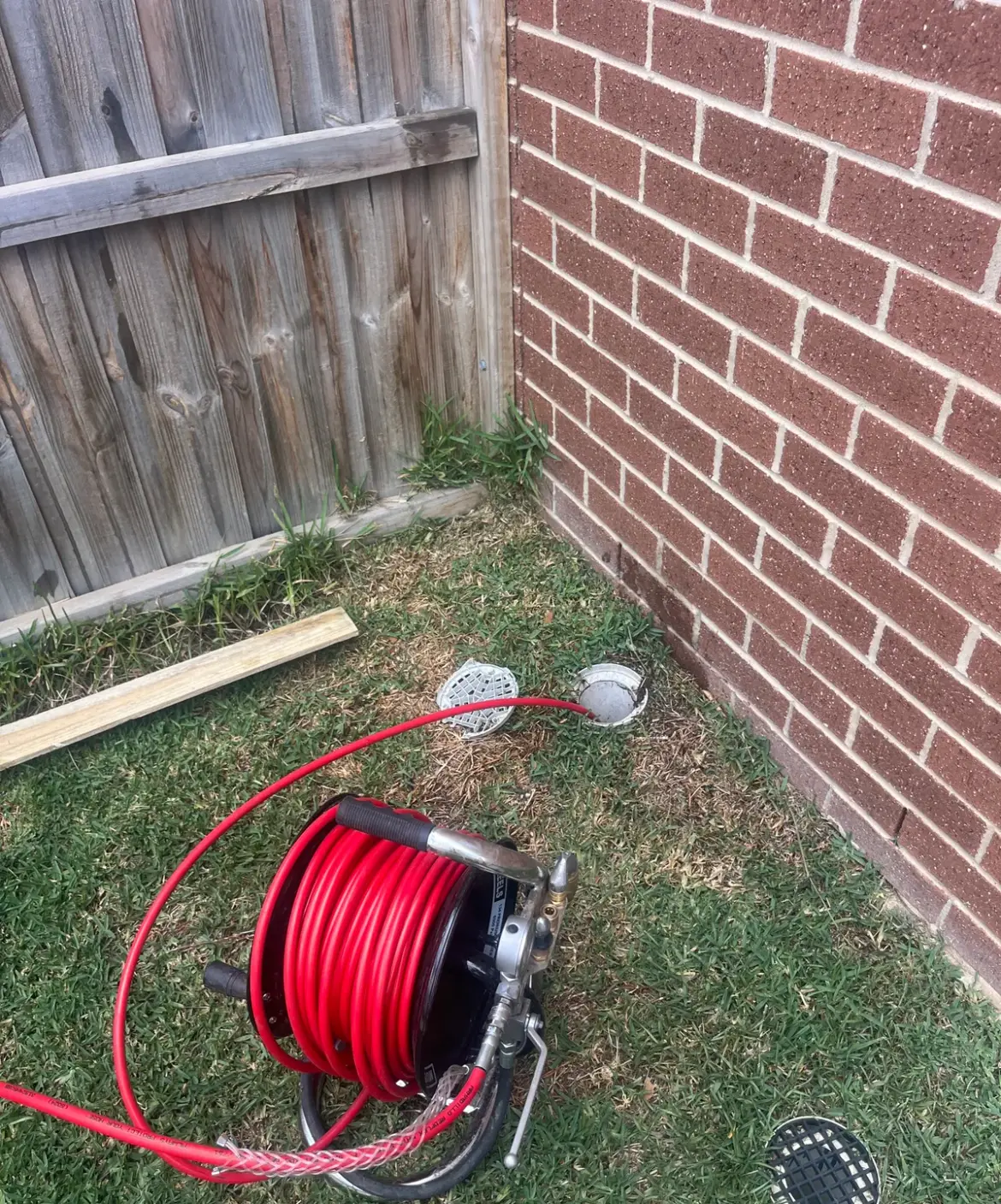 Red high-pressure hose reel on grass with the hose running into an open drain beside a brick wall and timber fence