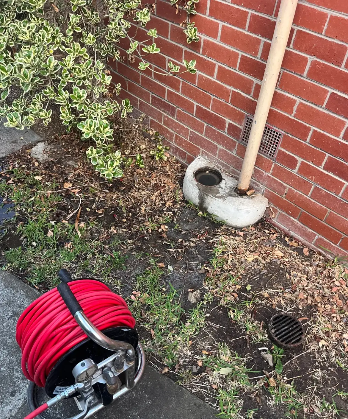 Red hose reel in front of a concrete-surrounded gully with a cream downpipe against a red brick wall