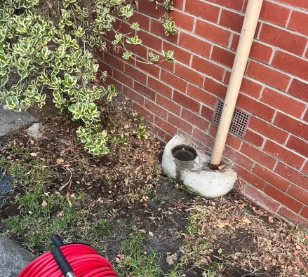 Red hose reel in front of a concrete-surrounded gully with a cream downpipe against a red brick wall