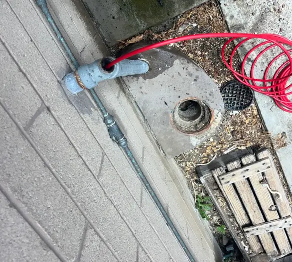 Red hose running into a grey pipe over a drain, beside a gully trap and timber slat cover