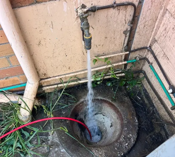 Red jetter hose in a drain pit with water flowing beside a brick wall and a hot water unit