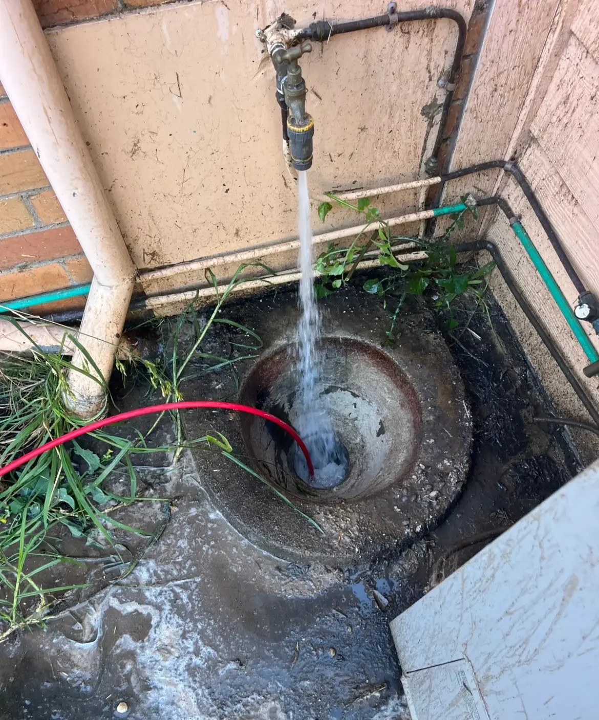 Red jetter hose in a drain pit with water flowing beside a brick wall and a hot water unit