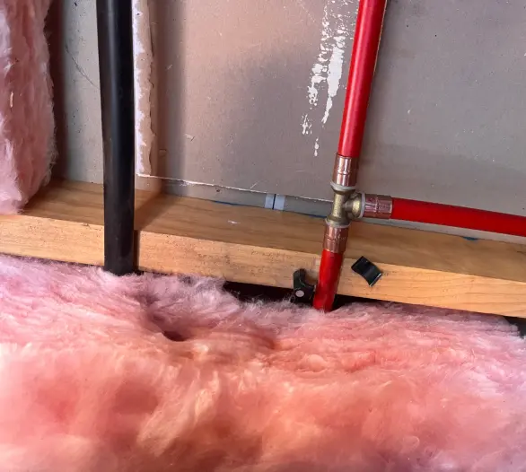 Red pipe and copper tee fittings inside an opened wall cavity beside pink insulation batts