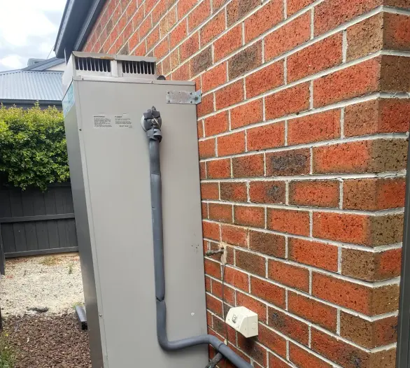 Tall grey hot water unit with insulated pipework beside a red brick wall
