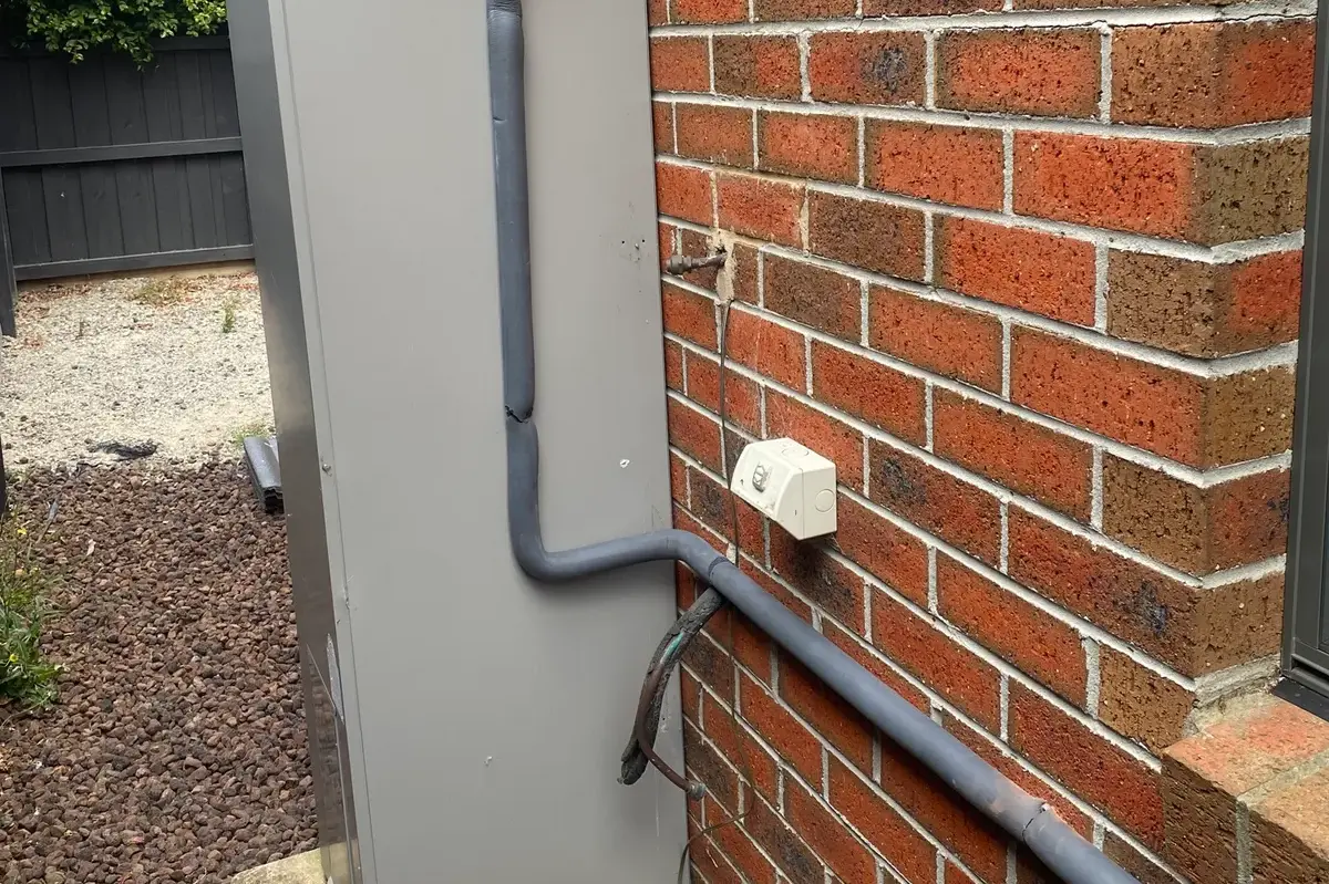 Tall grey hot water unit with insulated pipework beside a red brick wall