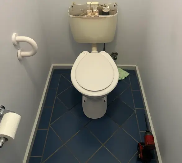 Toilet in a small room with the cistern lid off and a cordless drill on the floor