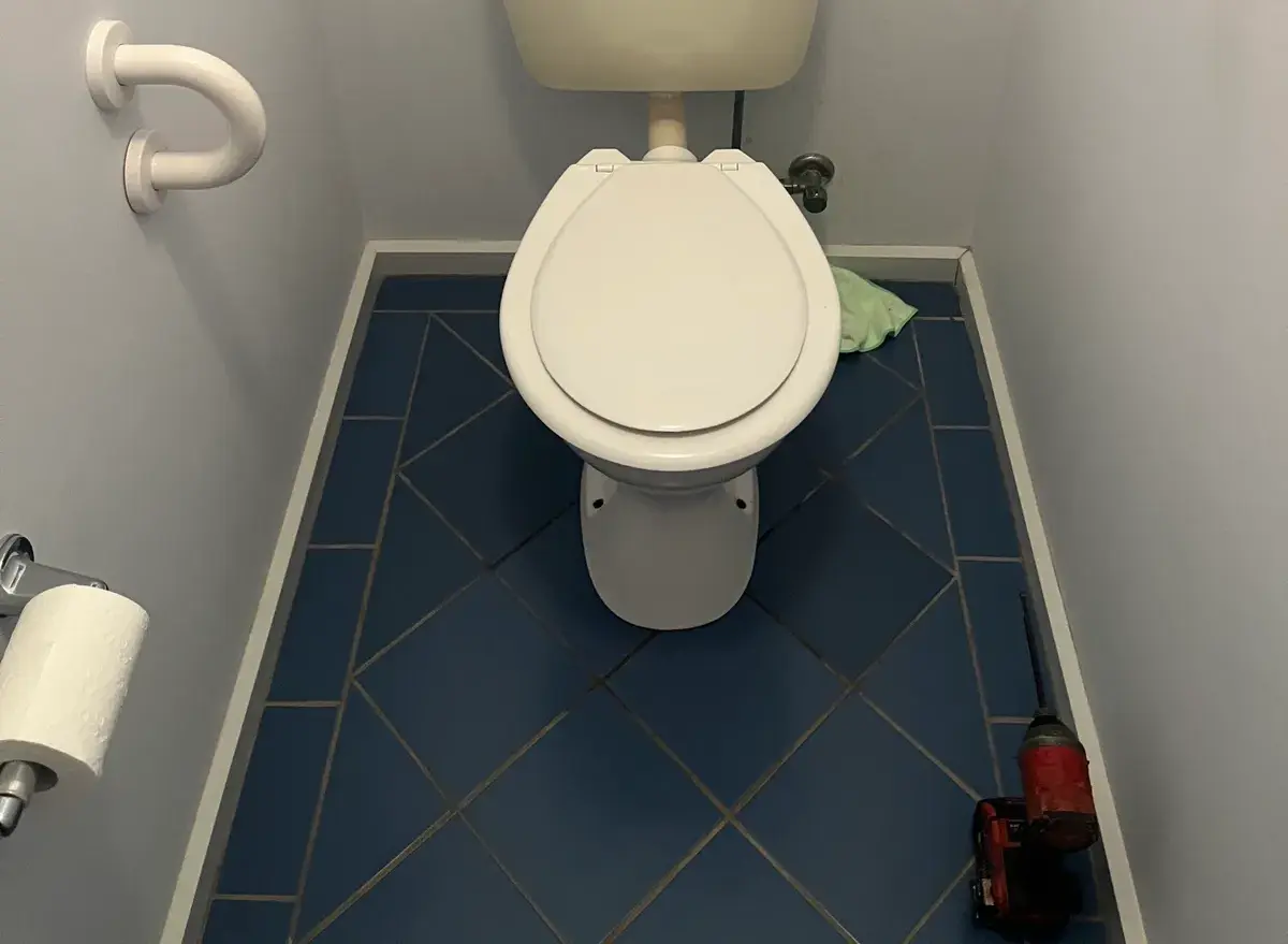 Toilet in a small room with the cistern lid off and a cordless drill on the floor