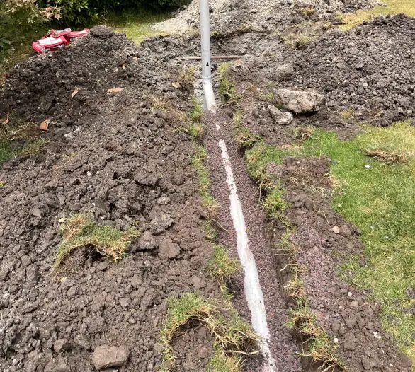 Trench dug in a backyard with new pipe laid below a fence and bushes