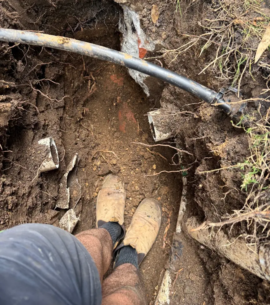 Trench dug in soil with a black pipe above and a worker looking down at their boots