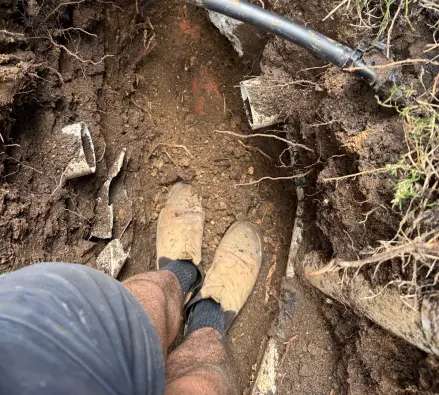 Trench dug in soil with a black pipe above and a worker looking down at their boots