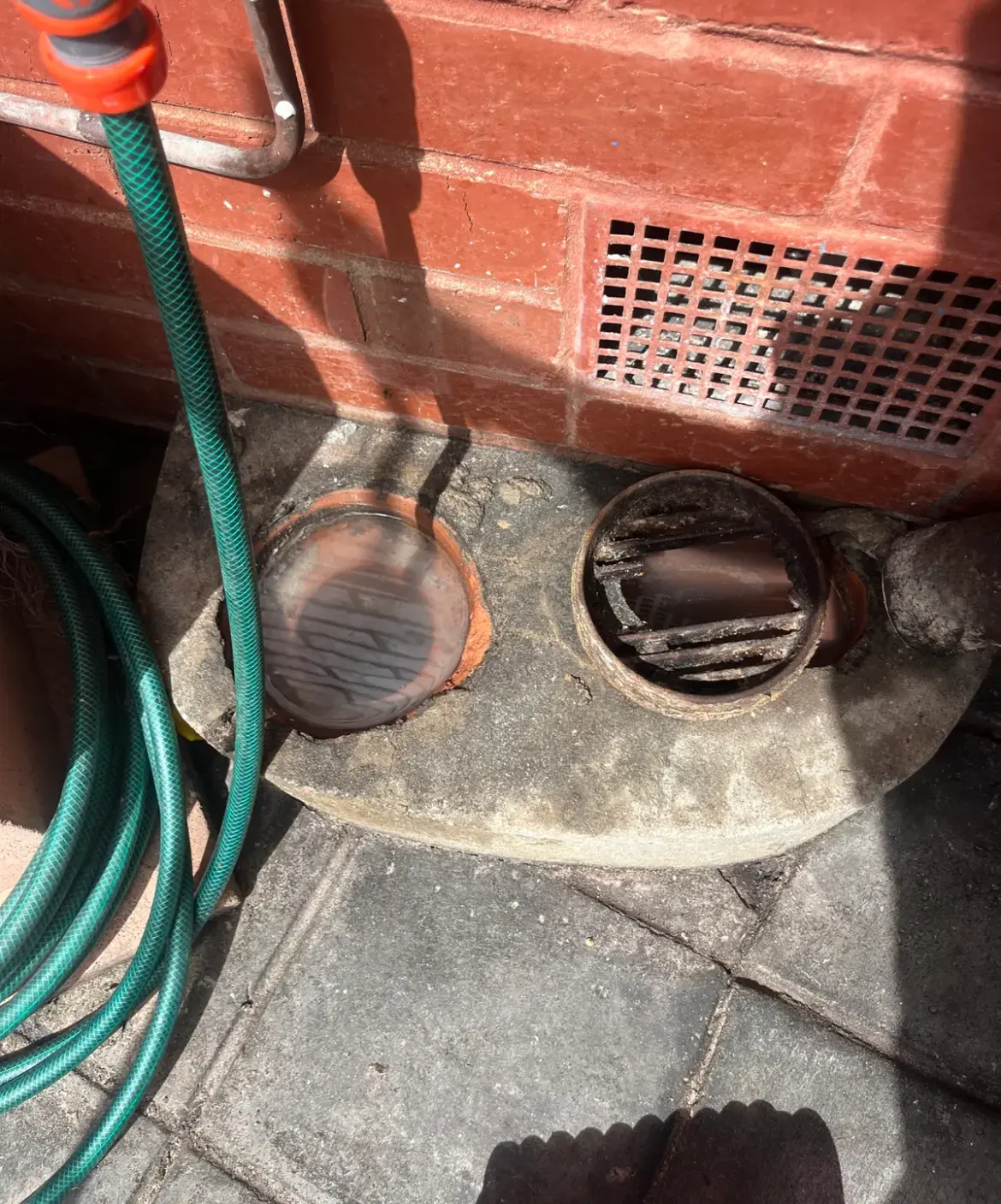Two round drain openings set in a concrete base against a brick wall, with a garden hose coiled beside them