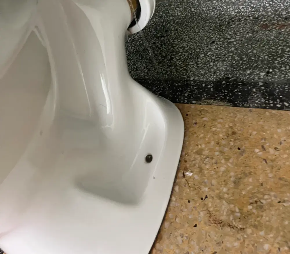 Underside of a toilet pan where it meets the pipe, with a thin stream of water and an isolation tap above