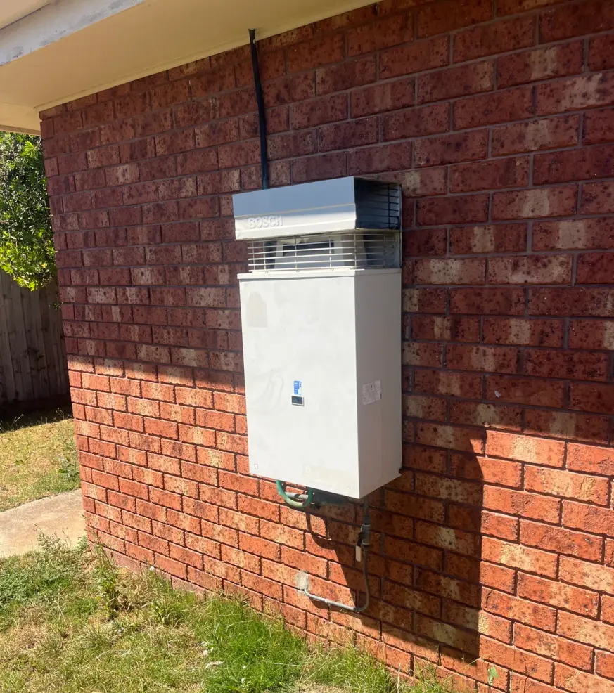 Wall-mounted continuous flow hot water unit on a brick wall beside a garden