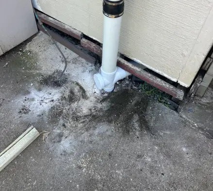 White pipe with a rubber coupling and junction at the foot of a weatherboard wall, stained concrete below