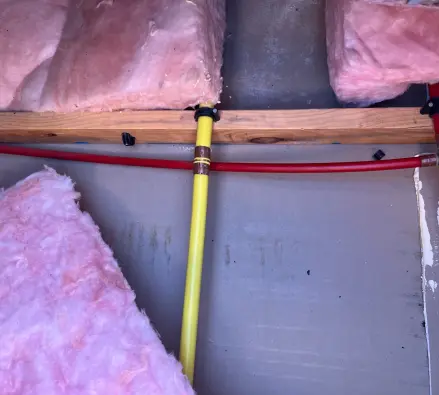 Yellow gas pipe and red water pipe running through an opened wall cavity with pink insulation batts